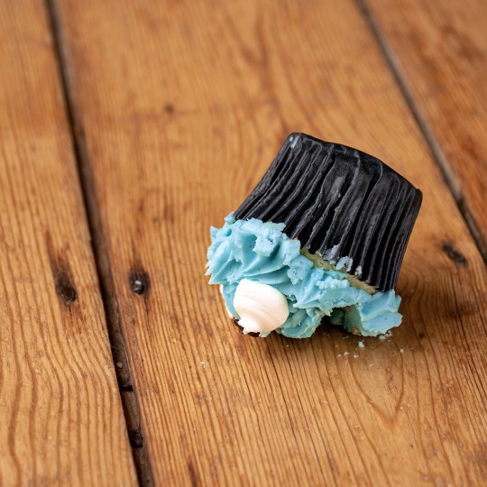 Ein Cupcake mit blauem Zuckerguss und schwarzer Verpackung liegt umgekippt auf einer Holzoberfläche.