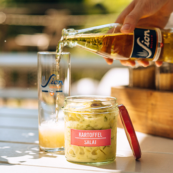 Eine Hand gießt Bier in ein Glas neben einem Glas Kartoffelsalat auf einem Tisch im Freien.