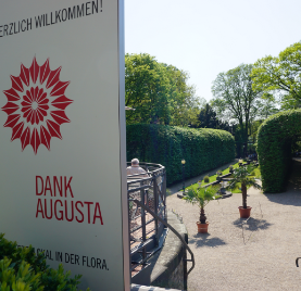 Ein Willkommensschild mit der Aufschrift „DANK AUGUSTA“ neben einem Gartenweg mit Topfpflanzen und Grünpflanzen.
