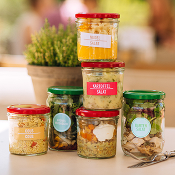 Auf einem Tisch sind sechs Gläser mit verschiedenen Salaten gestapelt, jedes mit der Sorte beschriftet, neben einer Topfpflanze.