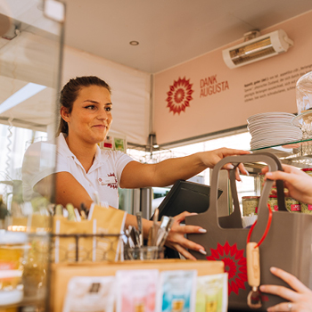 Eine lächelnde Frau überreicht einem Kunden an der Theke in einem hellen Café eine Einkaufstüte.
