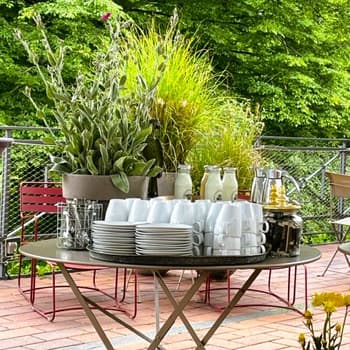 Runder Tisch mit gestapelten Tellern, Tassen, Gläsern und Krügen im Freien, umgeben von üppigem Grün.
