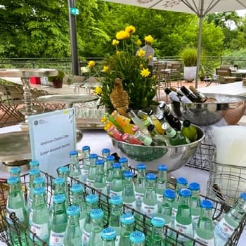 Tisch im Freien mit Getränkeflaschen, Metallschüsseln und gelben Blumen. Stühle und Grünpflanzen im Hintergrund.