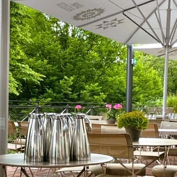 Mehrere Kaffeekannen aus Metall auf einem runden Tisch im Freien, mit Sitzgelegenheiten auf der Terrasse und viel Grün im Hintergrund.