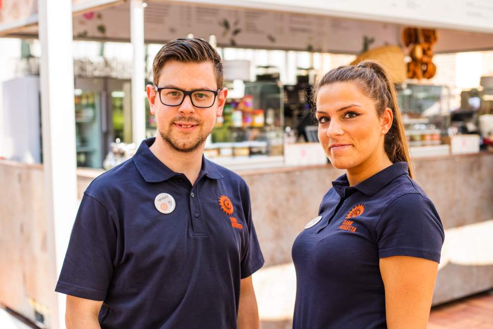 Zwei Mitarbeiter in marineblauen Poloshirts stehen lächelnd in einem hellen, modernen Café.