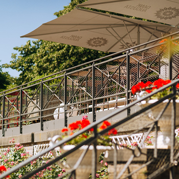 Caféterrasse im Freien mit Metallgeländer, roten Blumen und großen Sonnenschirmen an einem sonnigen Tag.